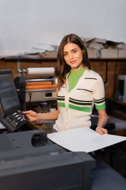 charming woman holding blank paper near printer in copy center  clipart