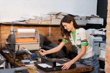 cheerful typographer using paper trimmer next to print plotter in print center  clipart