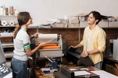 cheerful woman pulling paper while working with print plotter next to happy colleague   clipart