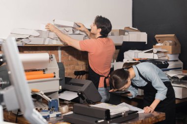 typographer in apron reaching folded carton boxes next to colleague and equipment in print center  clipart