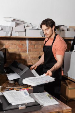 good looking typographer in apron holding printed newspapers in copy room  clipart