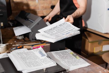 cropped view of typographer in apron holding printed newspapers with economic news  clipart