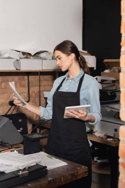 typographer in apron holding digital tablet and looking at newspaper in print center  clipart