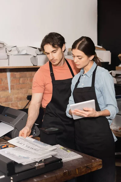 stock image typographer in apron holding digital tablet and looking at newspaper near colleague in print center 