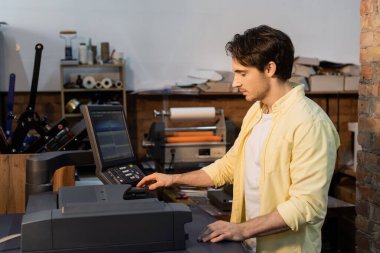 young man in yellow shirt using professional printer near monitor  clipart