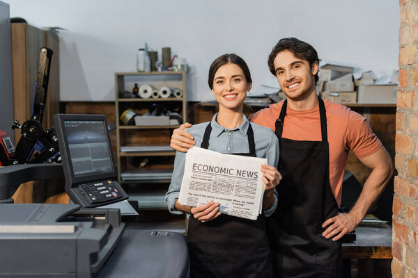 positive typographers in aprons smiling while holding printed newspapers with economic news 