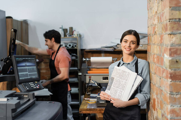 joyful typographer in apron holding newspapers near colleague on blurred background 