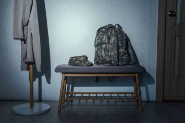 Military backpack and cap on bench in hallway at home at night  clipart