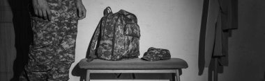 Black and white photo of soldier in uniform standing near backpack and car in hallway at home, banner  clipart