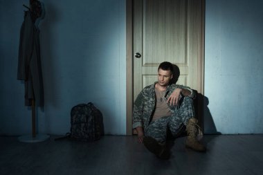 Hüsrana uğramış üniformalı bir asker gece koridorda, kapının yanında oturuyor. 