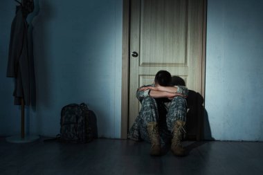 Lonely soldier in uniform sitting near backpack and door at home at night  clipart