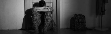 Black and white photo of depressed soldier sitting near backpack and door in hallway at home, banner  clipart