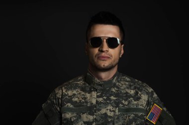 American soldier in camouflage uniform and sunglasses looking at camera isolated on black  clipart