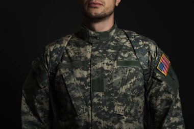 cropped view of patriotic American serviceman in military uniform with flag standing  isolated on black  clipart