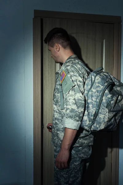 side view of American military man with backpack standing near entrance door at home