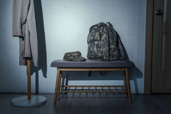 Military backpack and cap on bench in hallway at home at night 