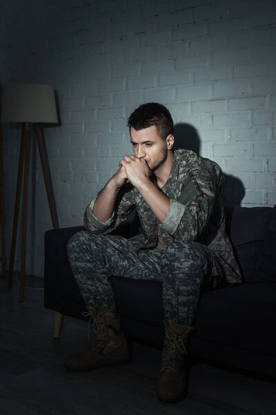 Frustrated soldier in camouflage sitting on couch and suffering from ptsd at home