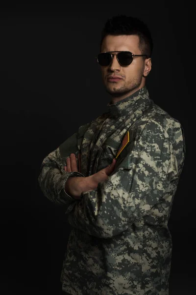soldier in camouflage uniform and sunglasses looking at camera while standing with folded arms isolated on black