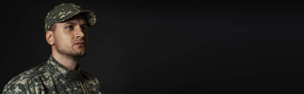 патриотический солдат в форме и кепке глядя в сторону изолированы на черный, знамя 