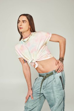 tattooed and young gay man with long hair standing in denim jeans and tied knot on t-shirt showing his belly and posing during pride month on grey background clipart