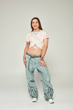 happy and tattooed young gay man with long hair standing in denim jeans and tied knot on t-shirt showing his belly and smiling during pride month on grey background clipart