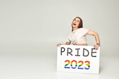 Heyecanlı, genç, eşcinsel, dövmeli, uzun saçlı, ağzı açık ve gri arka planda 2023 plaketi olan aktivist. Haziran 'da lgbt halk bayramı. 