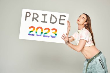 Dövmeli, uzun saçlı, kot pantolonlu ve tişörtünde düğüm olan genç eşcinsel bir adam. Gri arka planda gururlu 2023 plaketini tutarken göbeğini gösteriyor. 
