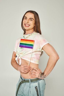 cheerful gay man with tattoo and long hair standing with hand on hip and bare belly while holding rainbow lgbt flag for pride month on grey background  clipart