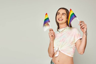 happy and tattooed homosexual man with long hair and bare belly smiling while holding rainbow lgbt flags for pride month on grey background  clipart