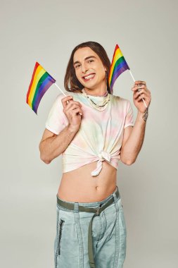 Uzun saçlı, çıplak göbekli, kot pantolonlu mutlu ve dövmeli bir gay gri arka planda gurur ayı için gökkuşağı bayrakları tutuyor. 