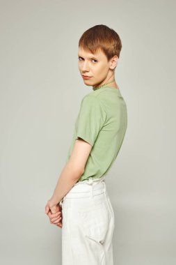 Gri arka plandaki gurur ayında kameraya bakarken beyaz kot pantolon ve yeşil tişörtle duran dudak parlatıcısı olan genç eşcinsel.