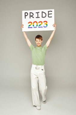 Dudak parlatıcısı beyaz kot pantolon ve yeşil t-shirt 'lü mutlu, genç, eşcinsel bir adam. Gri arka planda 2023 plaketi taşıyor.
