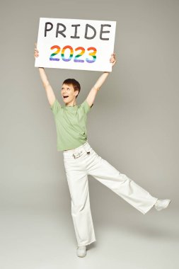 Dudak parlatıcısı ve beyaz kot pantolon ve yeşil tişörtle duran ağzı açık, neşeli, eşcinsel bir adam. Gri arkaplanda 2023 plaketi var.