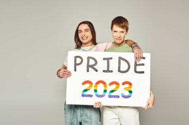 Igbt 'nin neşeli arkadaşları ellerinde 2023 plaketi tutuyorlar ve Haziran ayında stüdyoda gri arka planda lgbtq halk bayramını kutlarken kameraya bakıyorlar. 