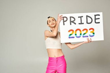 Beyzbol şapkalı, açık tişörtlü, pembe pantolonlu, gururlu 2023 plakalı genç ve mutlu bir adam. Gri arka planda durup kameraya bakıyor. 