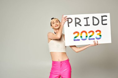 tattooed and pleased young gay man in baseball cap, crop top, and pink pants holding pride 2023 placard while standing and looking at camera on grey background  clipart