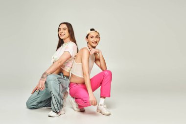 Pozitif ve dövmeli eşcinsel adam, gri arka planda oturup gurur gününü kutlarken beyzbol şapkalı eşcinsel arkadaşıyla gülümsüyor. 