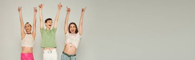 happy and young lgbt community friends in colorful clothes smiling while looking at camera, raising hands and celebrating pride month together on grey background in studio, banner  clipart