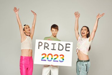 Mutlu ve dövmeli renkli giyinmiş eşcinsel adamlar elleri havada eşcinsel arkadaşının yanında ellerinde gururlu bir 2023 plaketi tutarken Haziran ayında lgbt halk bayramını kutluyorlar, gri arka plan, stüdyo 