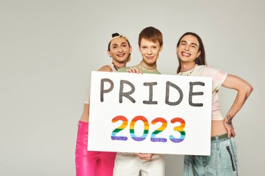 Çeşitli ve genç lgbtq topluluk arkadaşları. Renkli giysiler içinde duran ve gururlu 2023 plaketi tutan arkadaşları stüdyoda kameraya bakarken, gri arka plan. 