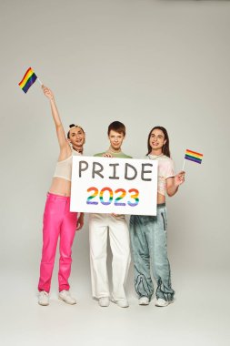 Neşeli ve dövmeli renkli giyinmiş eşcinsel adamlar ellerinde gökkuşağı bayraklarıyla eşcinsel arkadaşının yanında duruyor ellerinde gururlu bir 2023 plaketi var lgbt halk bayramını kutlarken, gri arka plan, stüdyo 