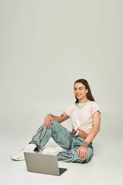 positive and young gay man with long hair and trendy clothes sitting and looking at modern laptop in studio on grey background during pride month  clipart