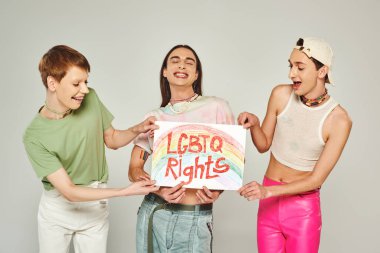 happy lgbt friends in colorful clothes holding placard with lgbtq rights lettering while standing together and smiling on pride month, grey background  clipart