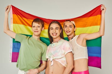 Renkli giysiler içinde bir grup mutlu gay ellerinde gökkuşağı bayraklarıyla gri arkaplanda gururlu bir arkaplanda kameraya bakarken yan yana duruyorlar.