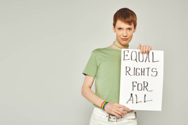 portrait of young queer activist wearing lgbt flag bracelet and holding placard with equal rights for all lettering looking at camera during pride month on grey background clipart