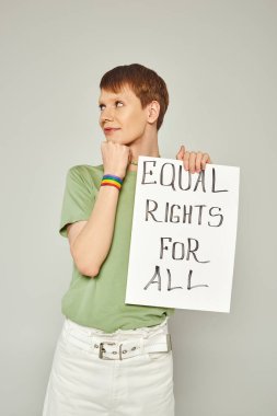 portrait of smiling queer activist wearing lgbt flag bracelet and holding placard with equal rights for all lettering looking away during pride month on grey background clipart