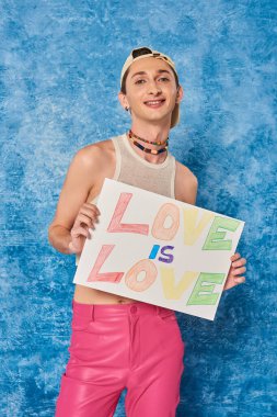 cheerful young gay activist in baseball cap and pink pants smiling while holding placard with love is love words during pride month on mottled blue background  clipart