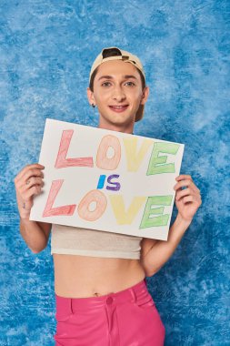 positive gay activist in baseball cap and white tank top smiling while holding placard with love is love words during pride month on mottled blue background  clipart