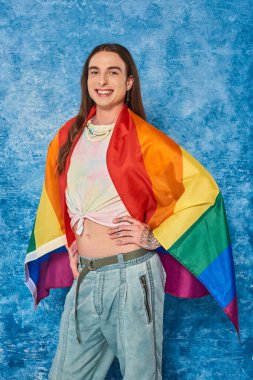 Uzun saçlı ve dövmeli mutlu gay adam. Günlük giysiler içinde Igbt bayrağıyla poz veriyor ve kameraya bakıyor. Gurur ayını mavi arka planda kutluyor.