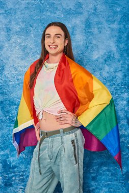 Happy long haired and tattooed gay man in casual clothes with lgbt flag posing and looking at camera while celebrating pride month on mottled blue background clipart